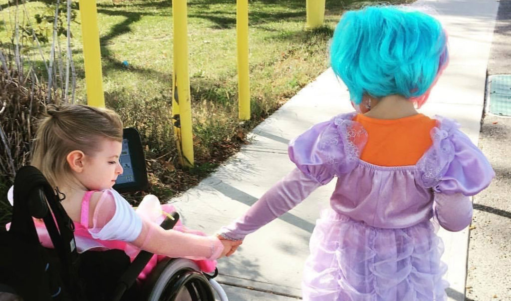 Two children in costumes holding hands, one using a wheelchair, walking together on a sidewalk.